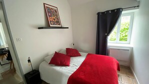 Ein Schlafzimmer mit einem Bett, roten Kissen, einer roten Decke, einem Wandregal mit einem Bild, einem Fenster mit Vorhängen und einem Heizkörper.