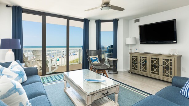 A spacious living room with a large glass door leading to a balcony overlooking a beach.