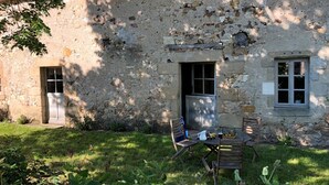 A rustic stone building with a wooden door and window, a small garden with a table and chairs, and a tree providing shade. - Restored Shepherd's Hut, Pets Allowed (Sauvagny)