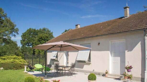 Une maison dotée d’un patio comprenant une table et des chaises sous un grand parasol, le tout entouré de plantes en pot et d’un jardin.