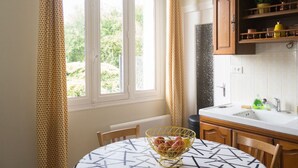 Une cuisine avec une table ronde, des chaises, un panier de fruits, une fenêtre avec des rideaux et des armoires en bois.
