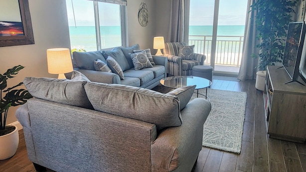 A living room with a grey sofa, a coffee table, and a TV stand. There is a balcony with a view of the ocean. - Oceanfront Corner 2BR Condo 2nd Floor Balcony Indian Harbour Beach, Unit 208 (Indian Harbour Beach)