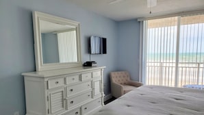 A bedroom with a large bed, a dresser with multiple drawers, a chair, a television on the wall, and a window with a view of the beach. - Oceanfront Corner 2BR Condo 2nd Floor Balcony Indian Harbour Beach, Unit 208 (Indian Harbour Beach)