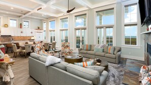 A spacious living area with a large gray sofa, a coffee table, and a dining table with chairs. There are large windows providing a view of the outdoors. - Huge Ocean View - Spa/Pool/Golf Cart (Port Aransas)