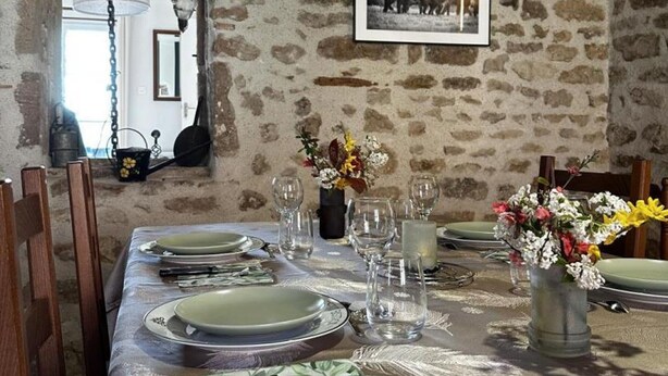 A rustic dining room with a stone wall, a framed picture, and a table set for four with plates, glasses, and a vase of flowers. - Country house, AC - 4 guests (Saint-Bonnet-Tronçais)