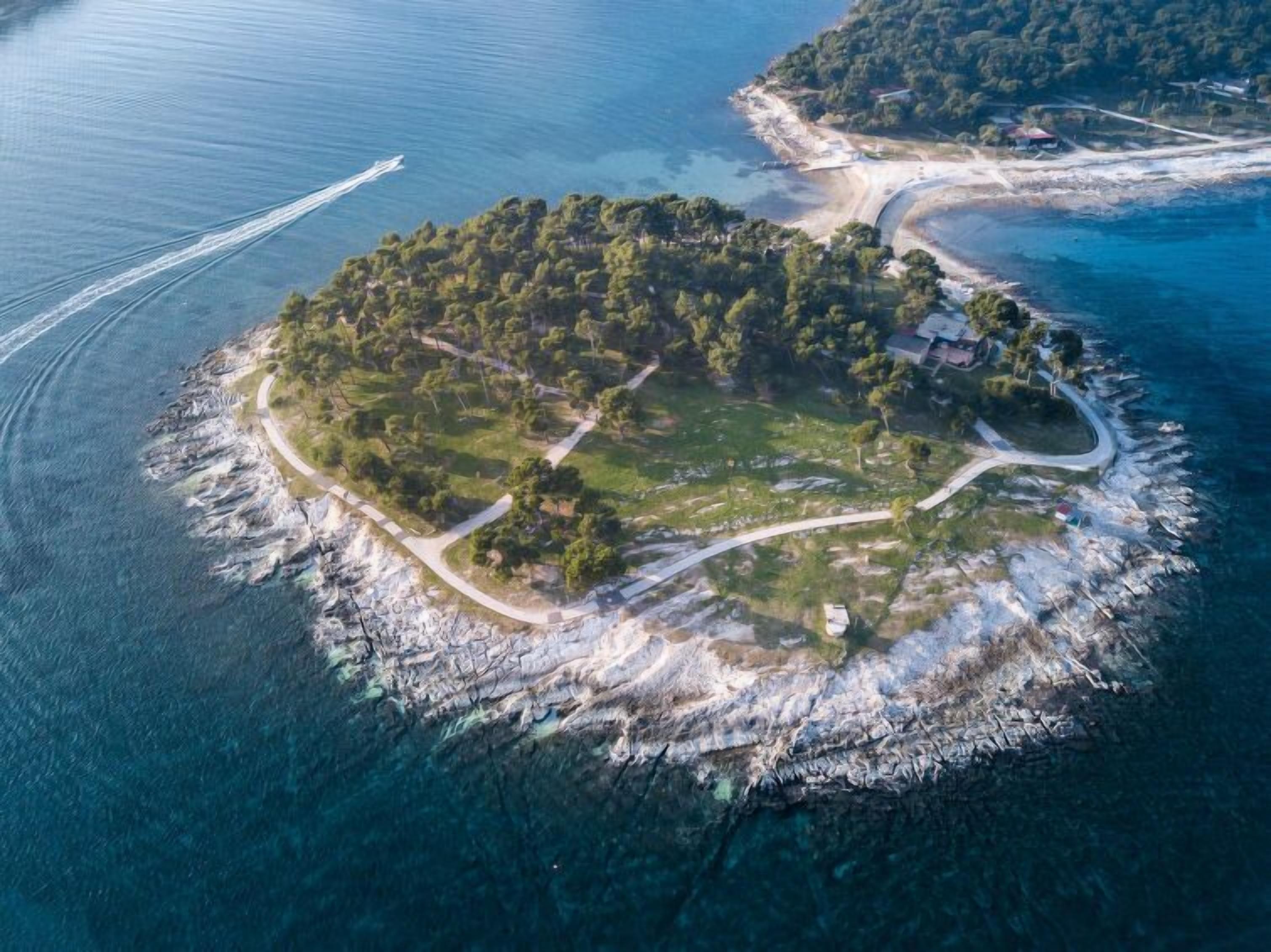 An aerial view of a small island with a winding road, surrounded by clear blue waters and dense greenery.