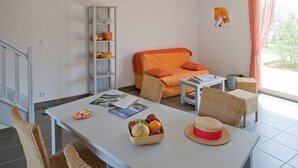 Una sala de estar con una mesa blanca, sillas de mimbre, un sofá con cojines naranjas, una estantería y una ventana con cortinas.