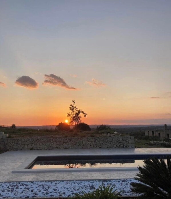 A sunset view over a swimming pool with a stone wall and a tree in the foreground.