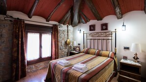 Un dormitorio con pared de piedra, vigas de madera en el techo, una cama con ropa de cama a rayas, una mesita de noche con una lámpara y una ventana con cortinas.
