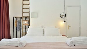 A neatly made bed with white linens and a pink blanket, a wooden ladder, a clothing rack with striped garments, and a round mirror on the wall. - La Perpetua - The Space of Mima (Noto)