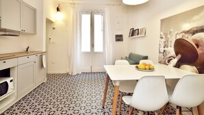 A compact kitchen with white cabinets, a black and white patterned floor, a dining table with chairs, and a fruit bowl. - La Perpetua - The Space of Mima (Noto)