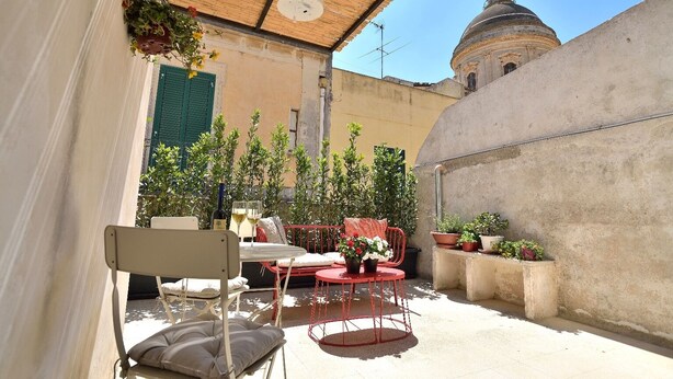 An outdoor patio with a table and chairs, surrounded by potted plants and a view of a historic building with a dome. - La Perpetua - The Space of Mima (Noto)