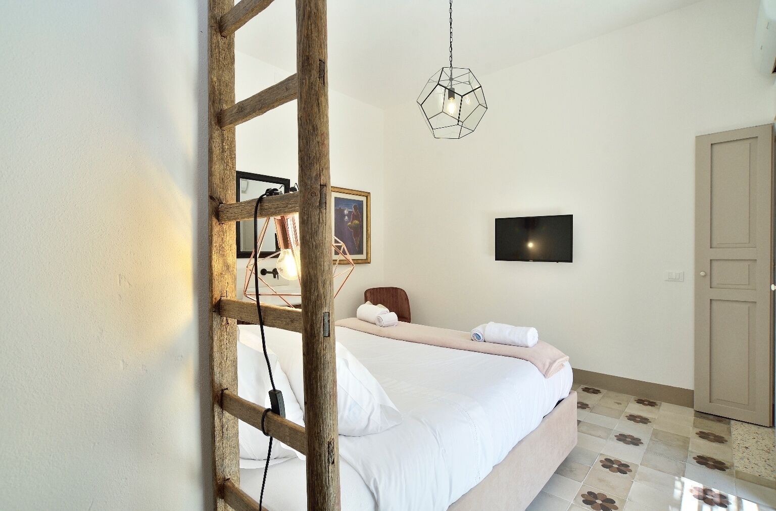A bedroom with a wooden ladder, a bed with white linens, a geometric pendant light, a framed picture on the wall, and a television.