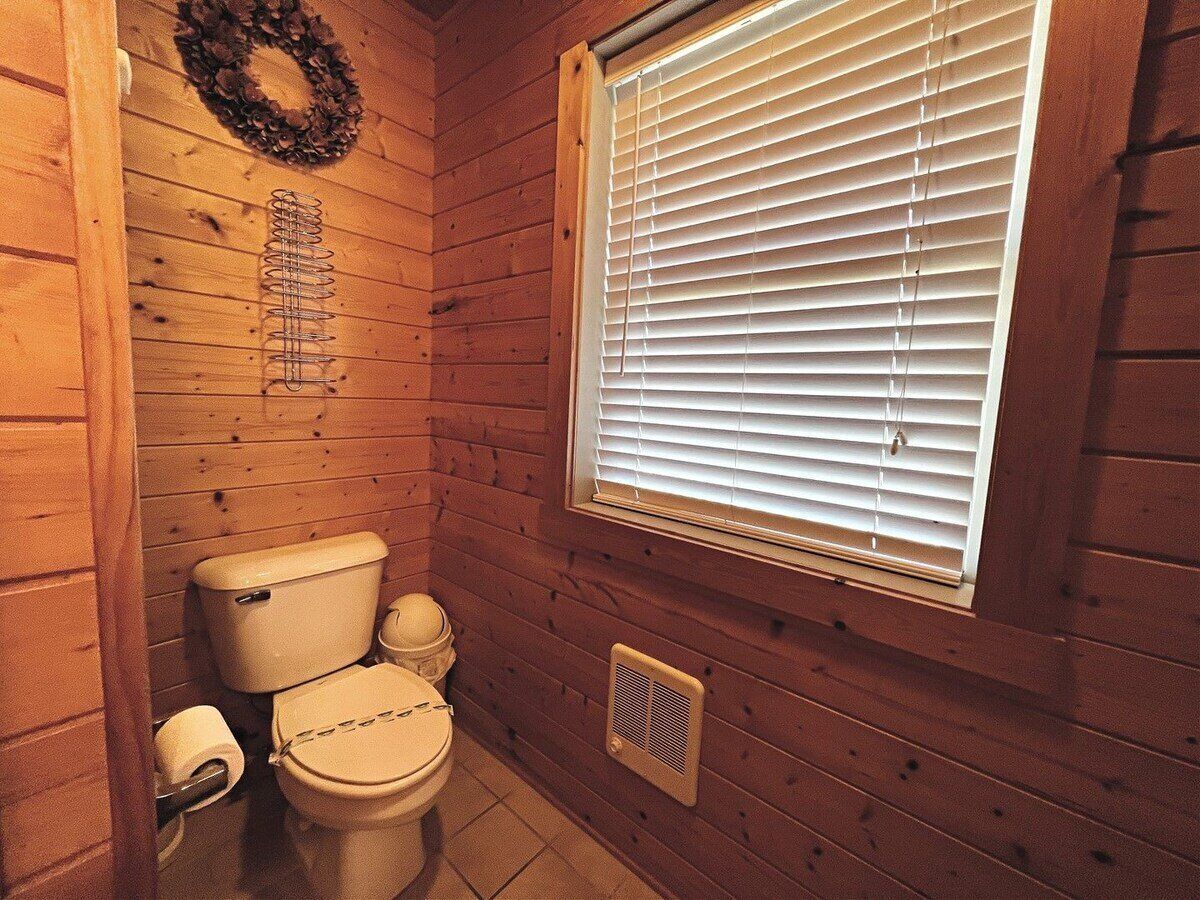 Een houten badkamer in een vakantiehuisje, met een toilet, een raam met zonwering, ventilatie en een krans aan de muur.