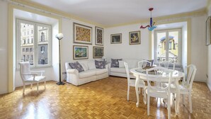 Un ampio soggiorno con un divano bianco, un tavolo da pranzo con sedie e una grande finestra che offre vista sugli edifici.