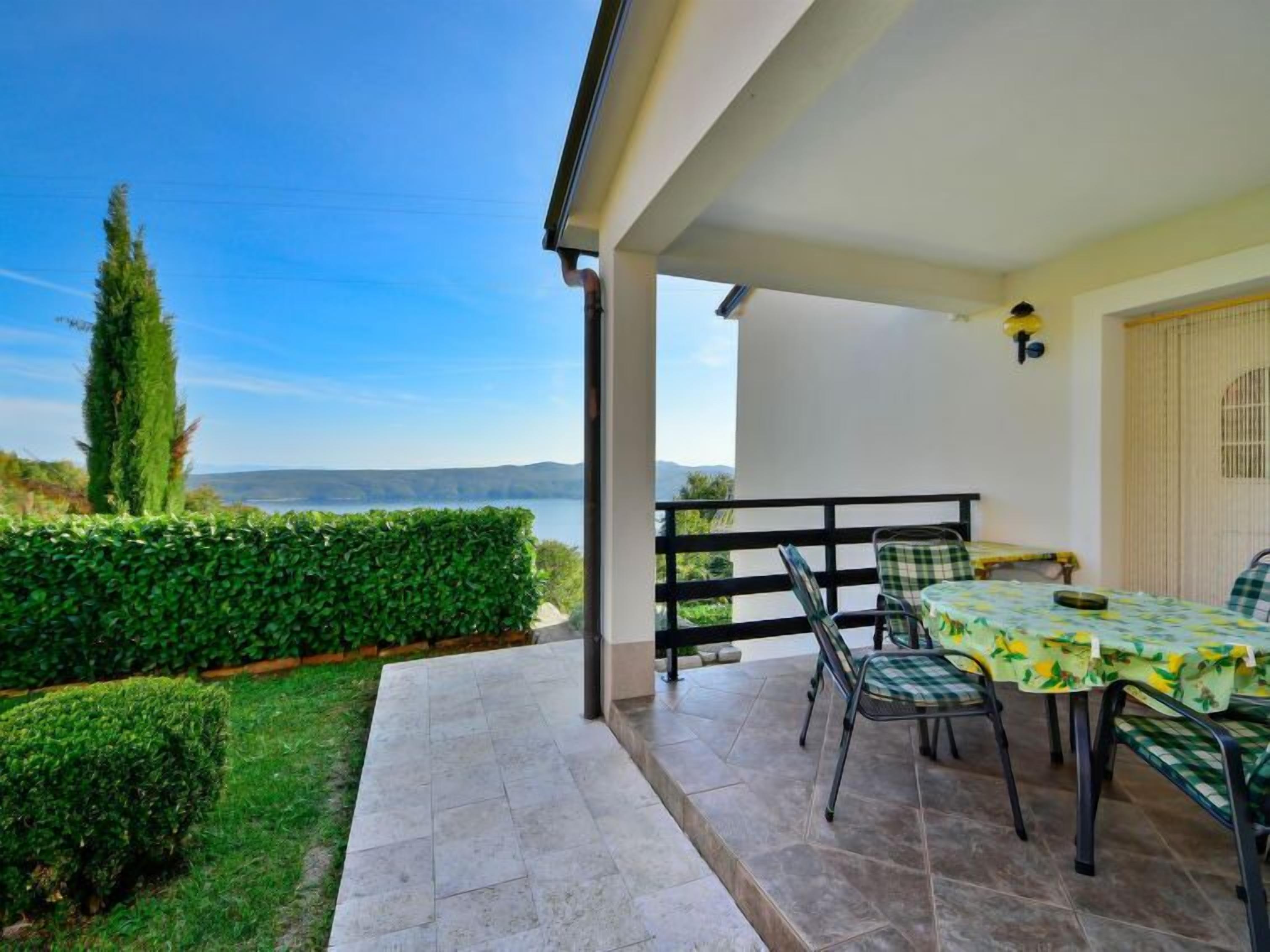 Eine überdachte Terrasse mit Tisch und Stühlen, Blick auf das Meer und ordentlich geschnittene Hecken.