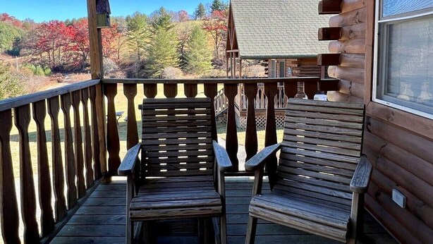 A wooden deck with two chairs and a table, overlooking a forested area with autumn foliage.