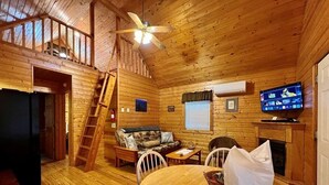 A wooden cabin interior with a dining area, a sofa, a television, and a fireplace.