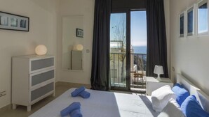 Ein Hotelzimmer mit Bett, Schminktisch, Nachttisch, Lampe und einem Balkon mit Meerblick.