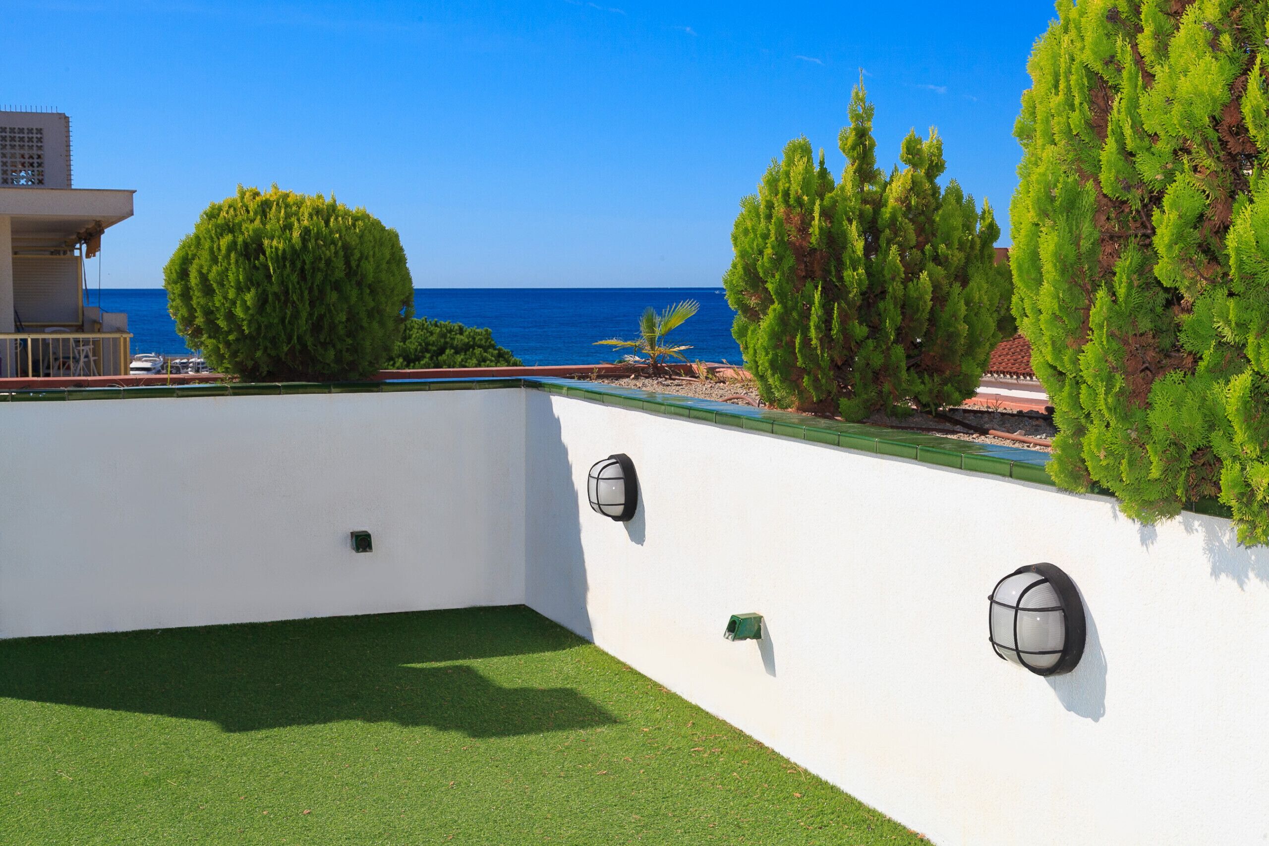 Une terrasse sur le toit offrant une vue sur la mer, recouverte d’un gazon synthétique vert, et un mur blanc agrémenté de deux appliques murales.