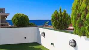 A rooftop terrace with a view of the sea, green artificial grass, and a white wall with two wall-mounted lights.