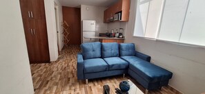 Ein kompakter Wohnraum mit einem blauen Sofa, einer Küchenzeile inklusive Kühlschrank und Mikrowelle sowie Holzboden.