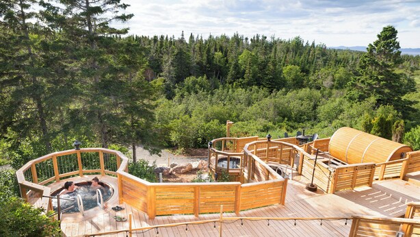A wooden deck with hot tubs and a wooden sauna, surrounded by a forest.