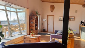 A spacious living room with a large window, wooden flooring, a ceiling fan, a wooden bookshelf, and a view of a snowy landscape.