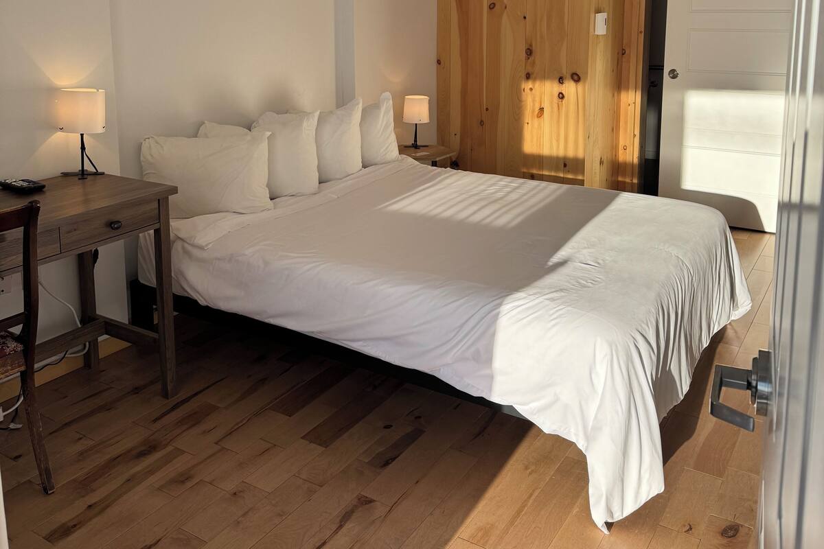 A bedroom with a wooden wardrobe, a bed with white bedding, a desk with a lamp, and a ceiling light.