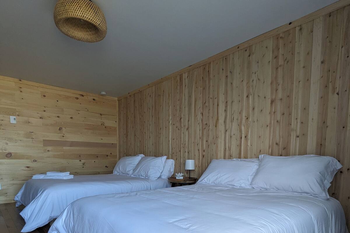 A room with two beds, wooden walls, and a woven ceiling light fixture.