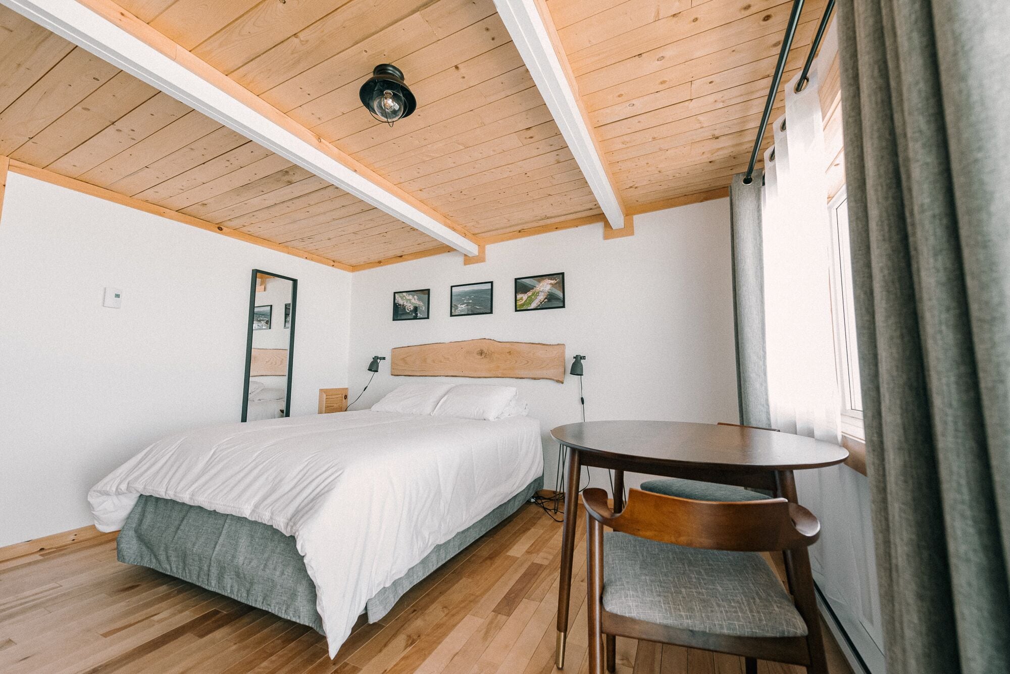 Quarto com teto de madeira, uma cama, mesa de cabeceira, uma cadeira e janela com cortinas.