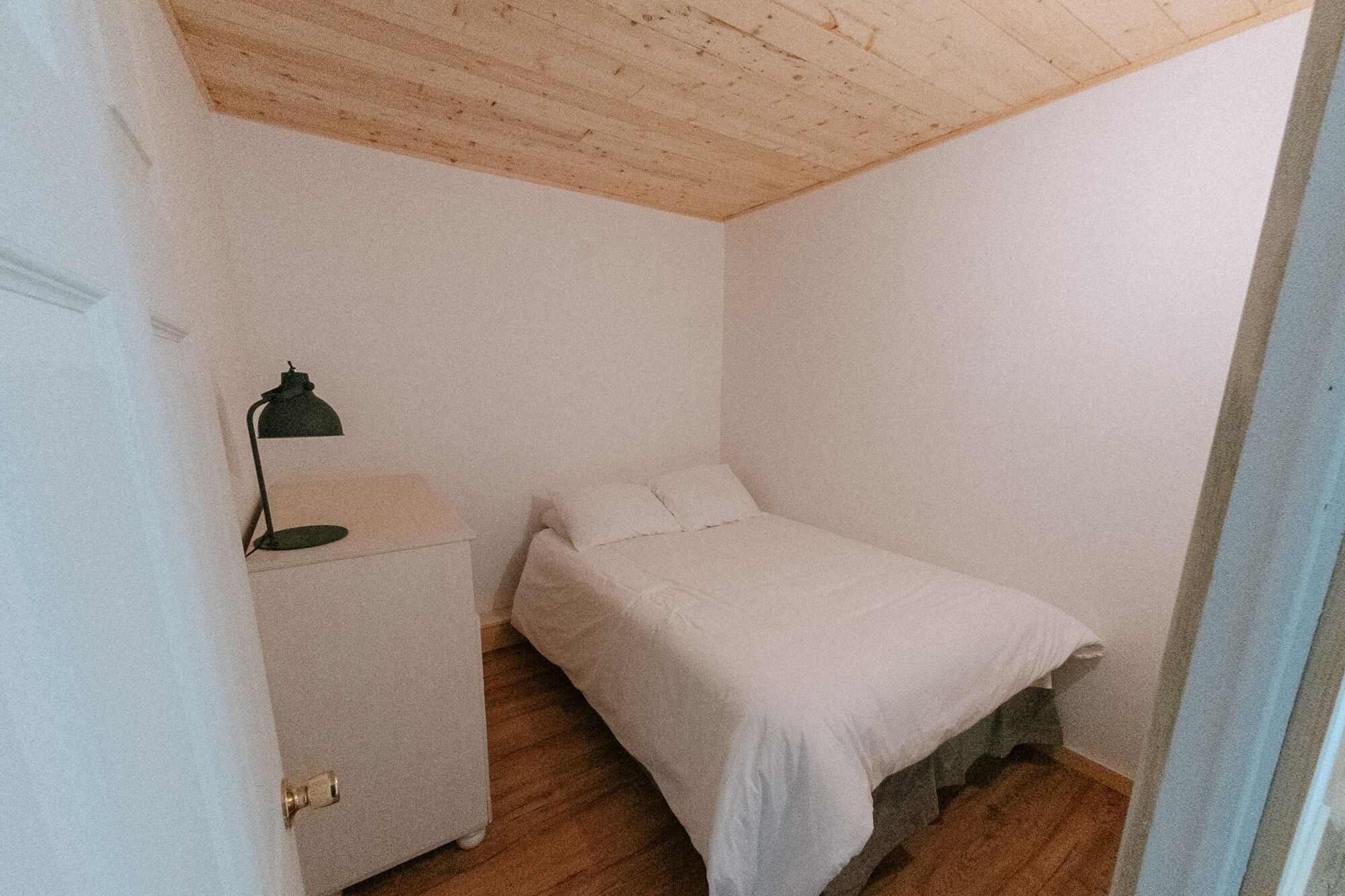 A small, sparsely furnished room with a wooden ceiling, a single bed, a nightstand, and a desk.