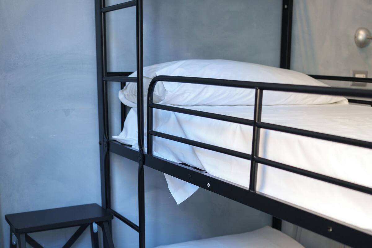 Una cama litera con ropa blanca, un taburete negro pequeño y una pared lisa.