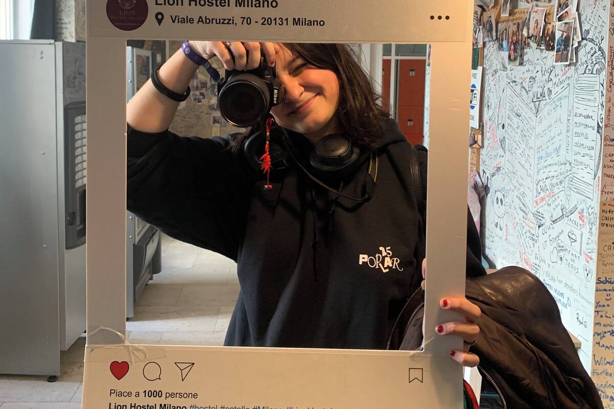 Un marco de cartón tipo Instagram con una foto de una persona sosteniendo una cámara, un hashtag para Lion Hostel Milano y una etiqueta de ubicación.