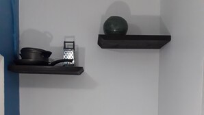A kitchen with two black shelves, one holding a grater and a pot, and the other a bowl. Below, there are various items including a basket, a box, glasses, and a mug. - Just Like Home, New Kingston (New Kingston)