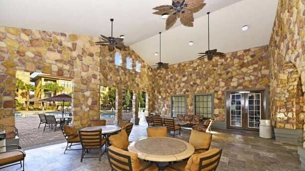 A stone-walled outdoor patio with round tables and chairs, ceiling fans, and a view of a pool and buildings in the background. - Bella Piazza 288924 (Davenport)