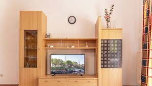 A living room with a TV, wooden cabinets, a clock, and a vase with flowers. - This bright holiday home on the ground floor offers on its approx. (Carolinensiel)
