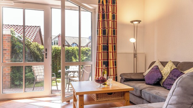 A living room with a sofa, a table with a bowl of fruit and a wine glass, a floor lamp, and large windows with a view of a garden and buildings. - This bright holiday home on the ground floor offers on its approx. (Carolinensiel)