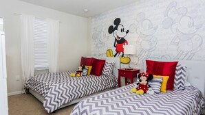 A child-friendly room with two beds, each with Mickey Mouse-themed bedding, a Mickey Mouse mural on the wall, and a Mickey Mouse plush toy on one bed. - Luxurious Vacation Townhome With Private Pool (Kissimmee)