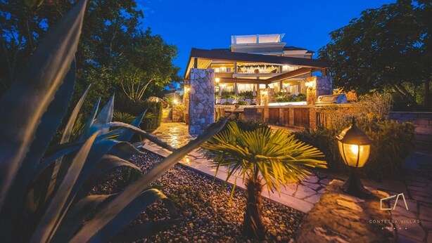 A modern house with a stone facade, a balcony with plants, and a well-lit outdoor area at dusk.