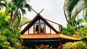 Een huis met een puntdak, omgeven door weelderige begroeiing en palmbomen.