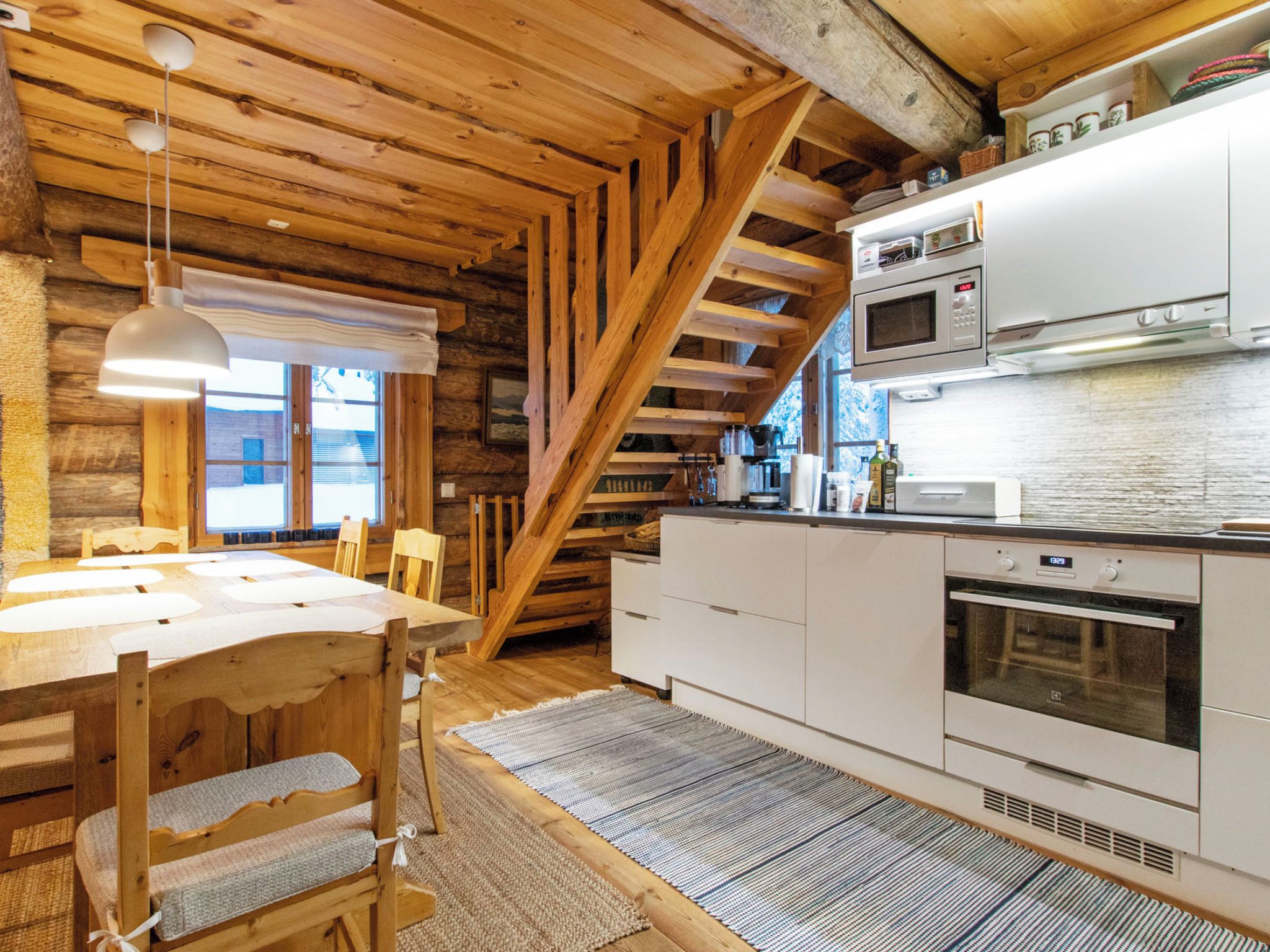 Interior rústico de una cabaña con una mesa de comedor de madera, sillas, una cocina con electrodomésticos blancos y un techo inclinado.