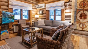A cozy log cabin interior with a sofa, chairs, a table, and a TV displaying a mountain scene.