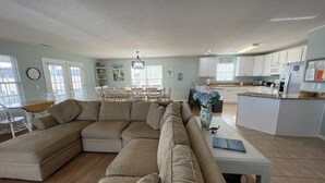 Amplia sala de estar con un sofá seccional beige grande, una zona de comedor con mesa y sillas de madera, y una cocina con armarios y electrodomésticos blancos.
