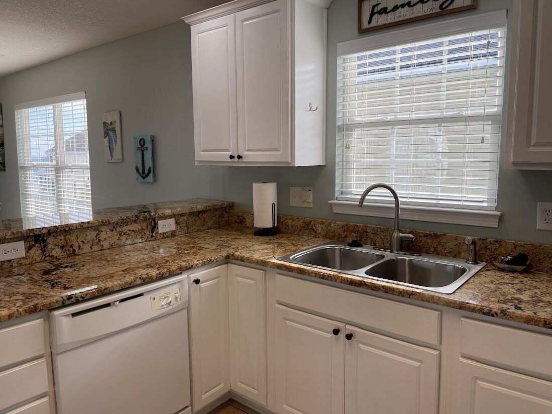 Een keuken met witte kastjes, granieten aanrecht, een dubbele spoelbak en een raam met zonwering.
