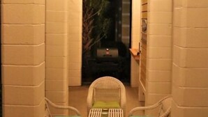 Un espacio de patio cubierto con dos sillas de mimbre y una mesa, rodeado de muros de ladrillo blanco y vigas de madera.