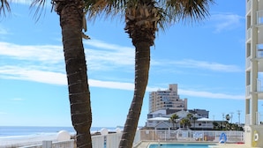 A pool area with palm trees, a beach view, and a high-rise building in the background. - One Ocean Place 1104 - Oceanfront Condominium in Garden City Beach (Garden City)