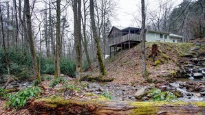 Una cabaña rústica con terraza de madera, rodeada de un bosque con un arroyo.