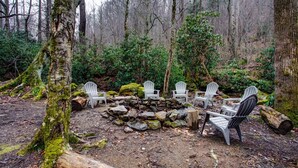 Un entorno forestal con un hogar de piedra y sillas Adirondack blancas dispuestas alrededor.