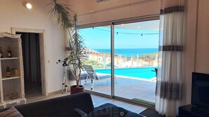 A living room with a sofa, a potted plant, and a view of a swimming pool and ocean.
